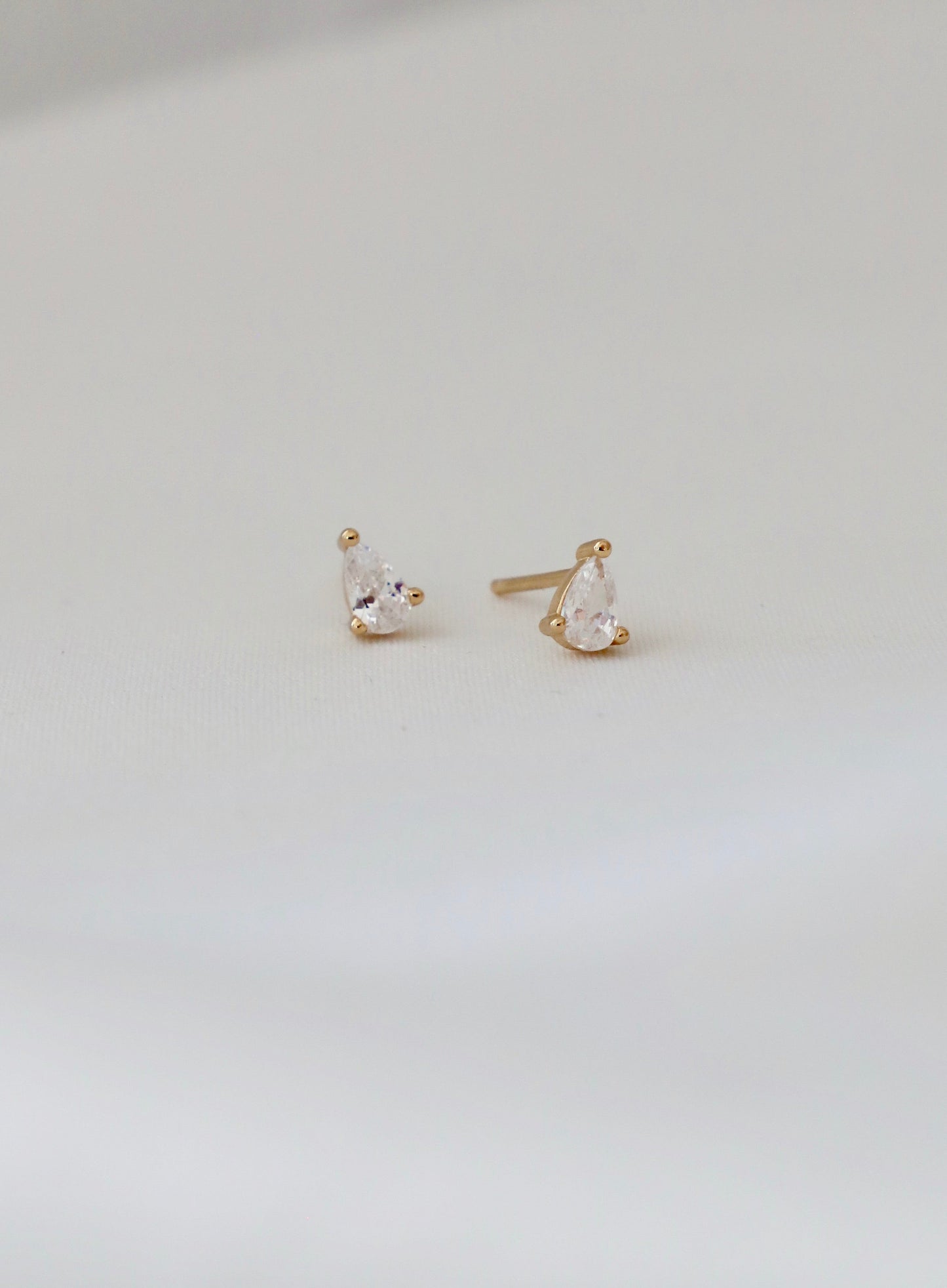 Two golden stud earrings, featuring pear-shaped clear gemstones, rest on a soft white surface, showcasing their elegant design and subtle shine in a well-lit context.