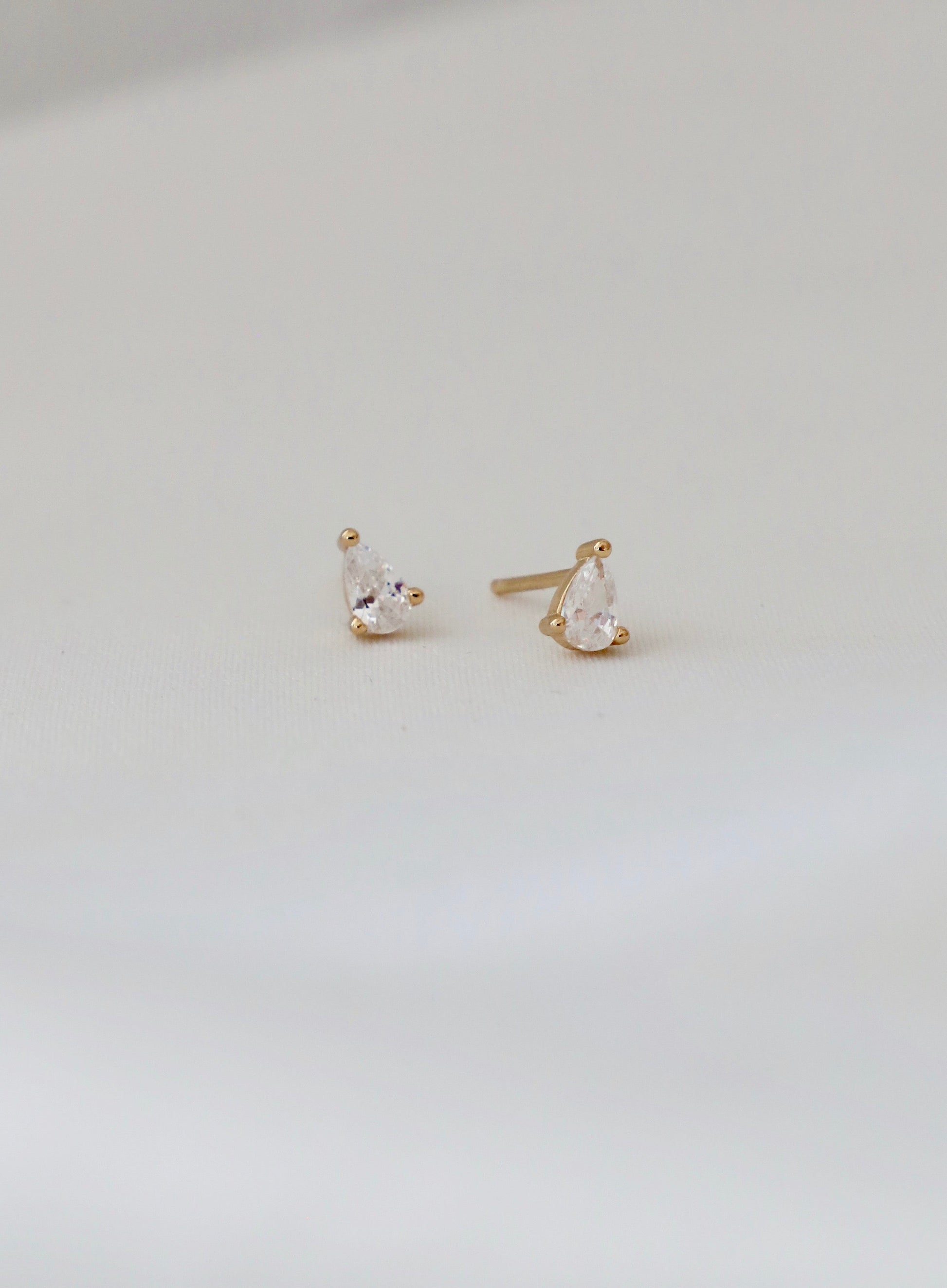 Two golden stud earrings, featuring pear-shaped clear gemstones, rest on a soft white surface, showcasing their elegant design and subtle shine in a well-lit context.