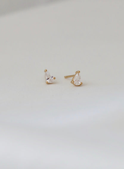 Two golden stud earrings, featuring pear-shaped clear gemstones, rest on a soft white surface, showcasing their elegant design and subtle shine in a well-lit context.