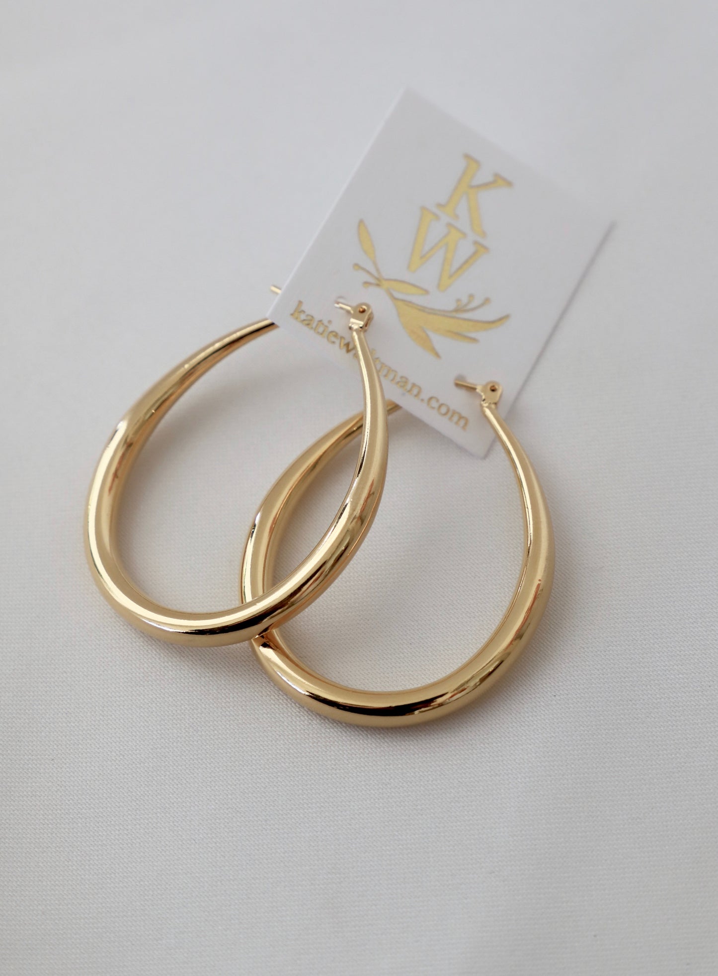 Gold hoop earrings rest on a light-colored fabric backdrop, showcasing their shiny, smooth surface. A small product tag featuring a logo and website is attached to the earrings.