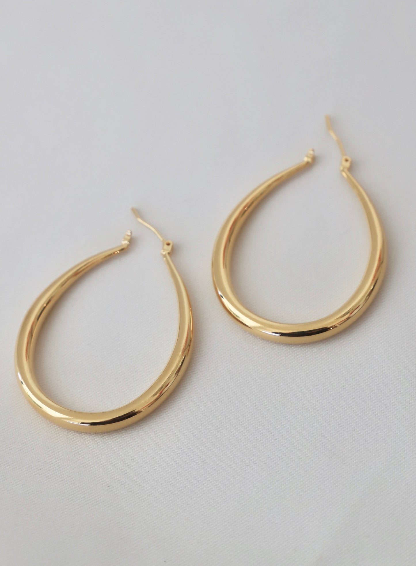 Two gold hoop earrings are displayed gracefully on a light-colored, textured fabric background, showcasing their shiny, smooth curves and elegant design.