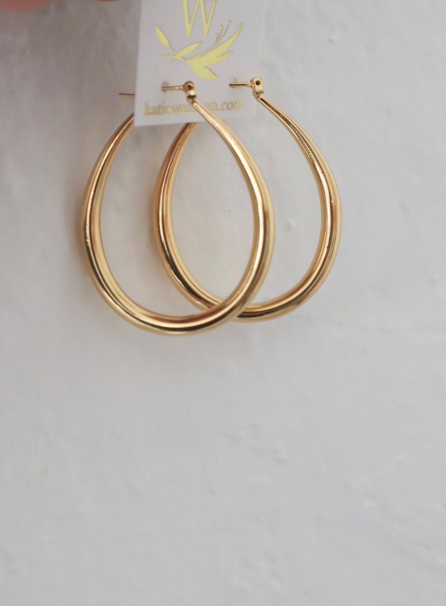 Gold hoop earrings hang from a display tag. They are shiny and have a slender, elongated shape, set against a textured white background.