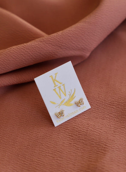 Gold butterfly-shaped earrings are displayed on a white card, resting on a textured, rust-colored fabric. The card features the initials "KW" and a website link below.