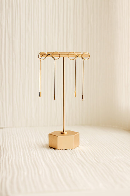 A gold earring stand displays three pairs of earrings, showcasing their elegant designs. The stand is set against a textured, light-colored background, enhancing the overall aesthetic.