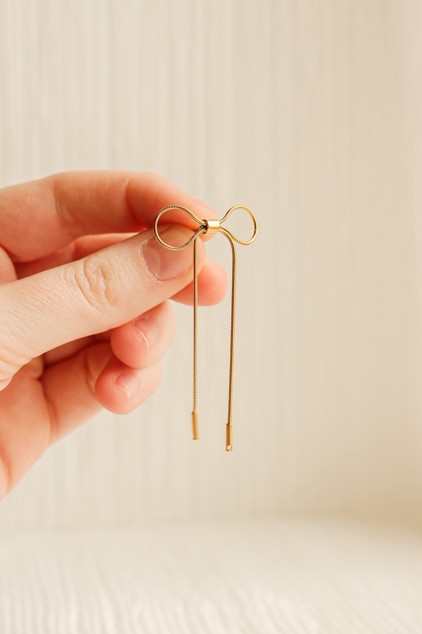 A hand holds a delicate gold bow-shaped object with two thin, hanging strands. The background features a lightly textured, neutral-toned surface, enhancing the elegance of the object.