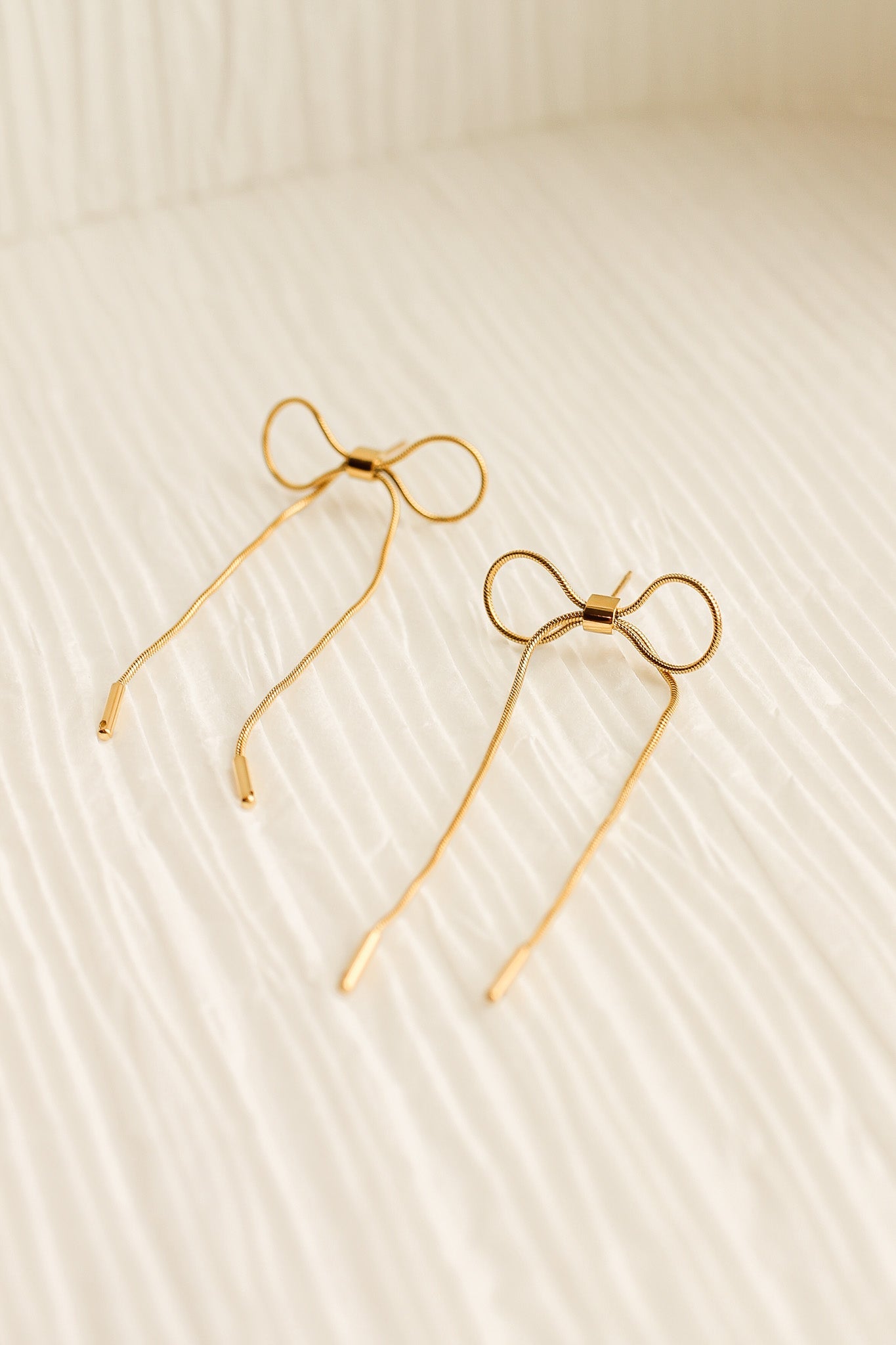 Two golden hairpins shaped like bows rest on a textured, light-colored fabric surface, creating a delicate and elegant appearance in a softly lit environment.