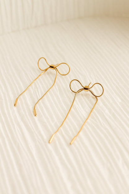 Two golden hairpins shaped like bows rest on a textured, light-colored fabric surface, creating a delicate and elegant appearance in a softly lit environment.