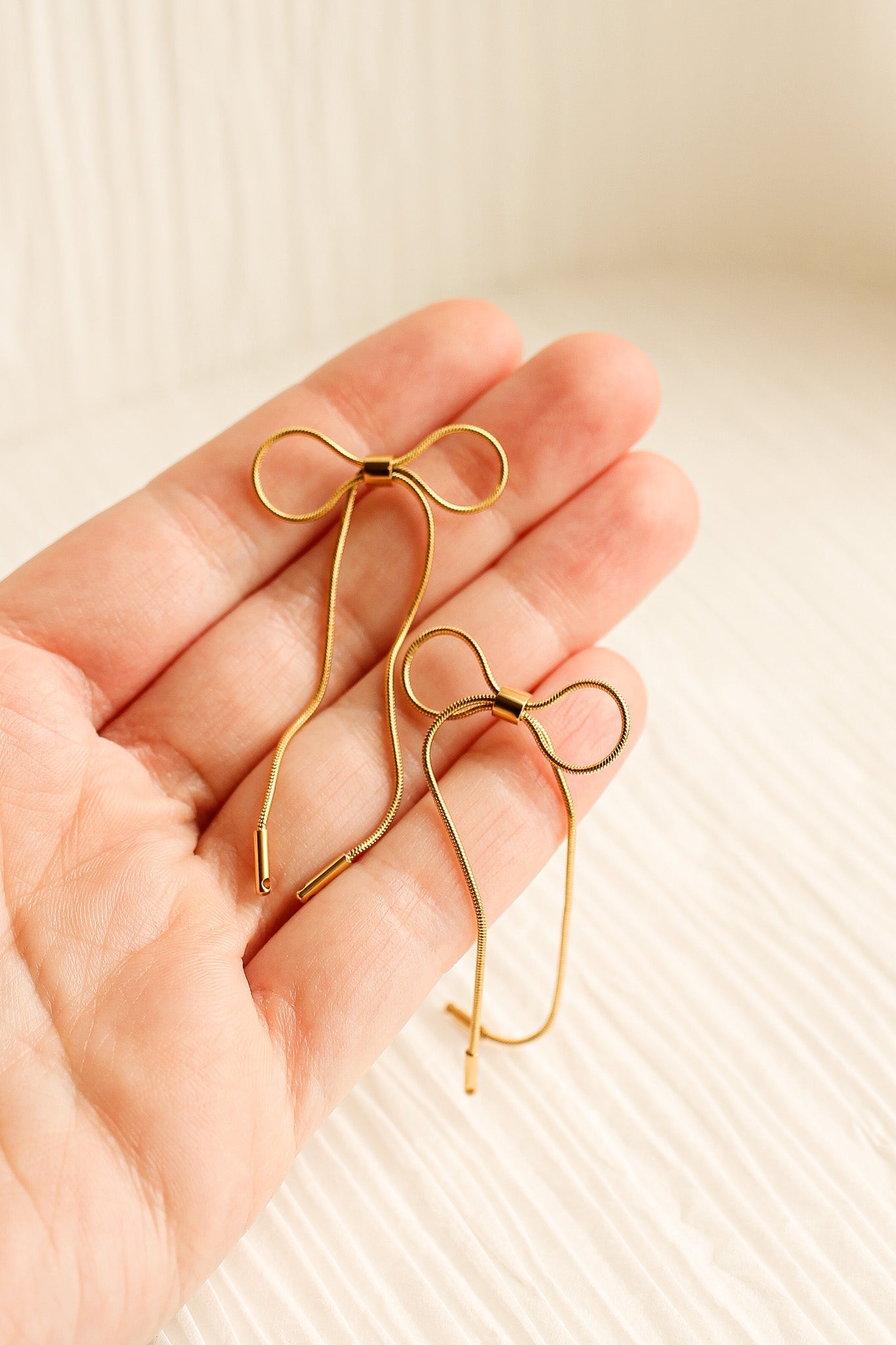 Two gold hairpins shaped like bows rest in an open palm. The background features a light, textured surface, suggesting a soft, neutral environment.