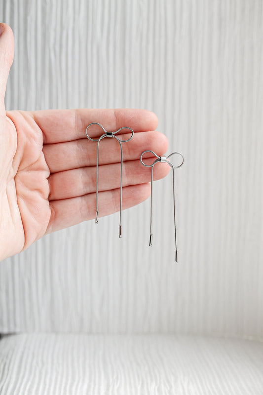 Two metal hairpins resembling bows are held in an outstretched hand. The background features a textured, light-colored surface, providing a simple and clean context.