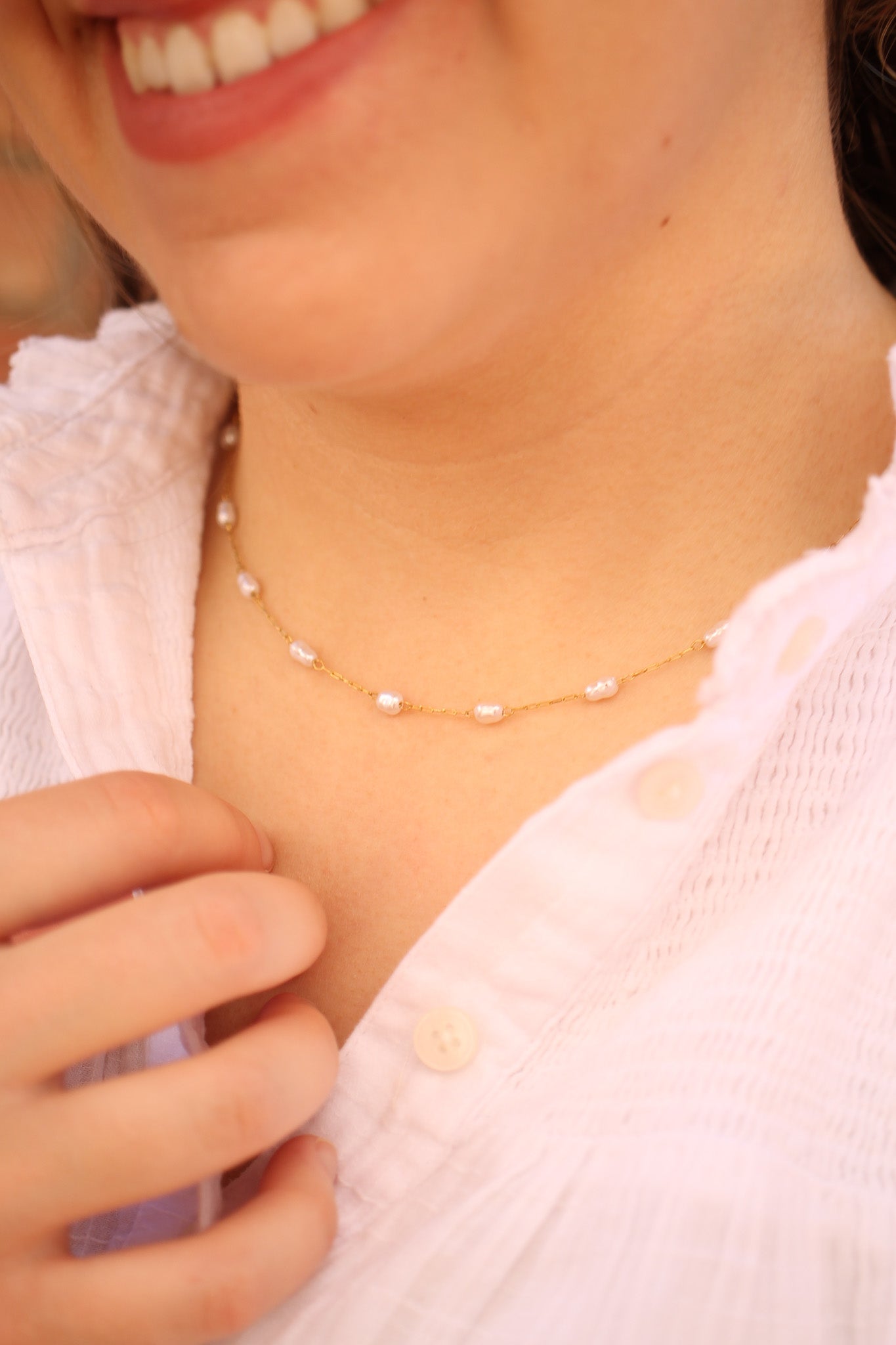 A necklace made of small pearls on a delicate gold chain is worn around a person's neck. The individual gently touches the necklace while wearing a light, button-up blouse.