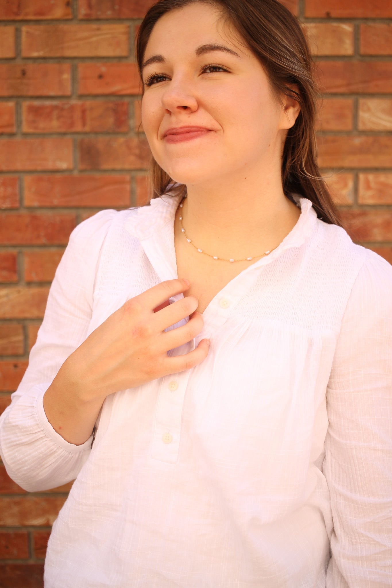 A young woman in a white blouse smiles softly while touching a delicate necklace around her neck, standing against a backdrop of red brick.