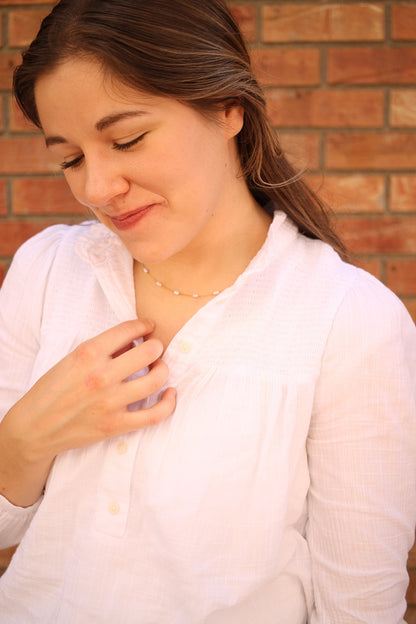 A young woman in a white shirt lightly touches a delicate necklace around her neck, smiling softly with her eyes closed, against a backdrop of brick wall.