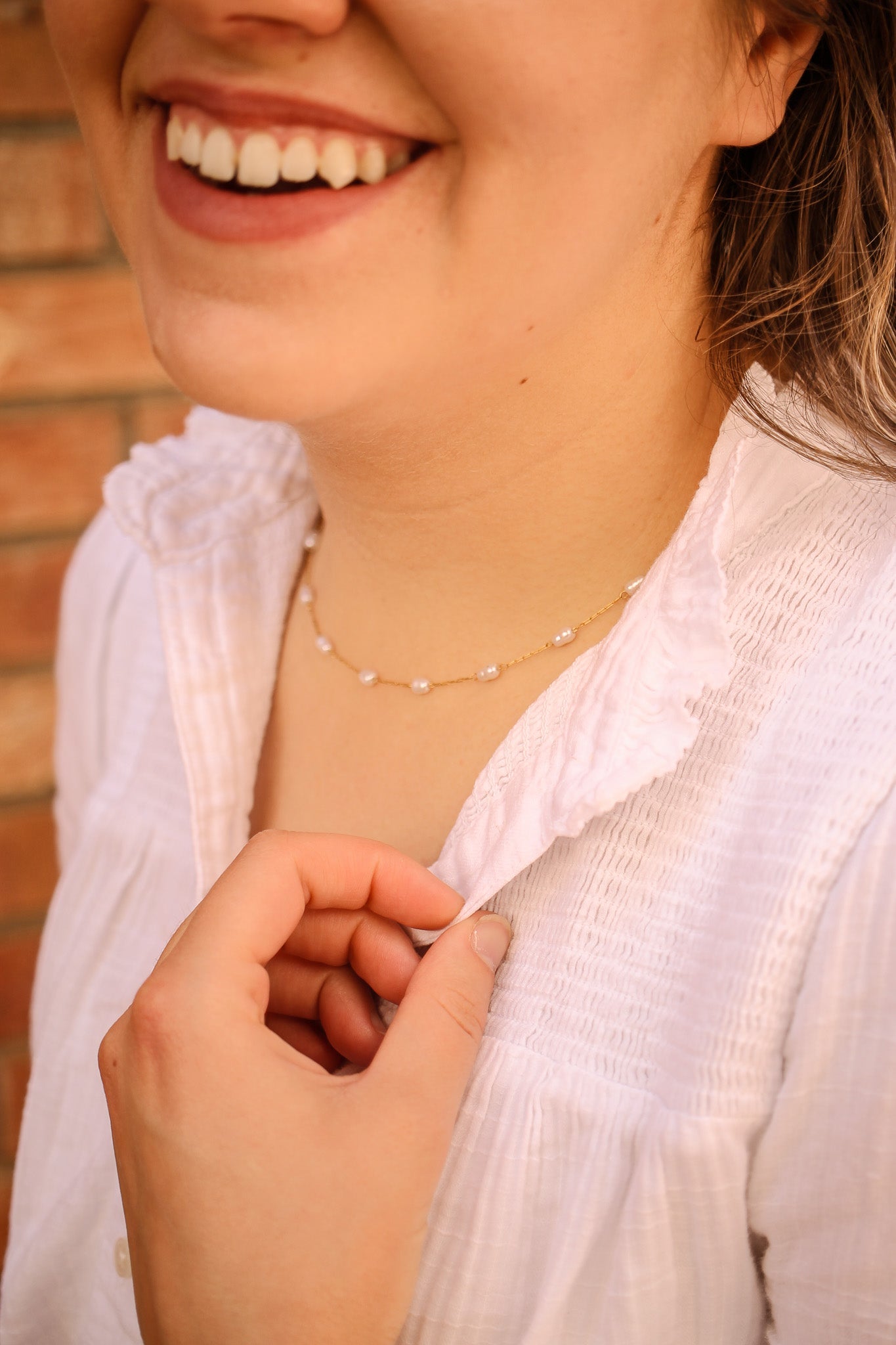 A woman smiles while touching the collar of her white shirt. She wears a delicate pearl necklace, and a brick wall provides a warm, textured background.