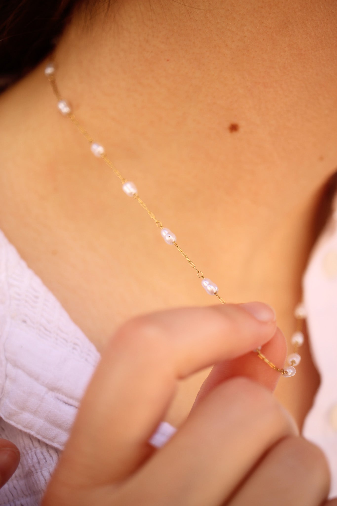 A hand gently holds a delicate gold chain adorned with small beads, resting against the skin of a neck. The background suggests a soft focus, enhancing the jewelry's elegance.