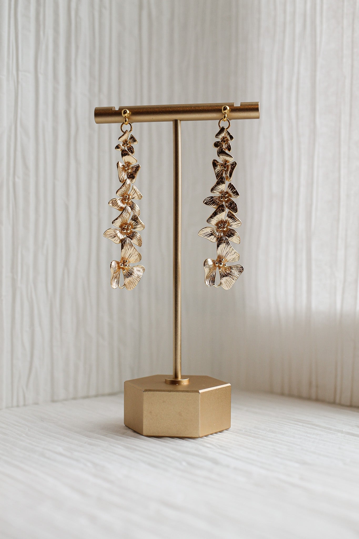 A pair of ornate, gold-colored earrings hangs from a vertical stand, showcasing their intricate floral design. The stand rests on a textured, light-colored surface, with soft, natural light illuminating the scene.