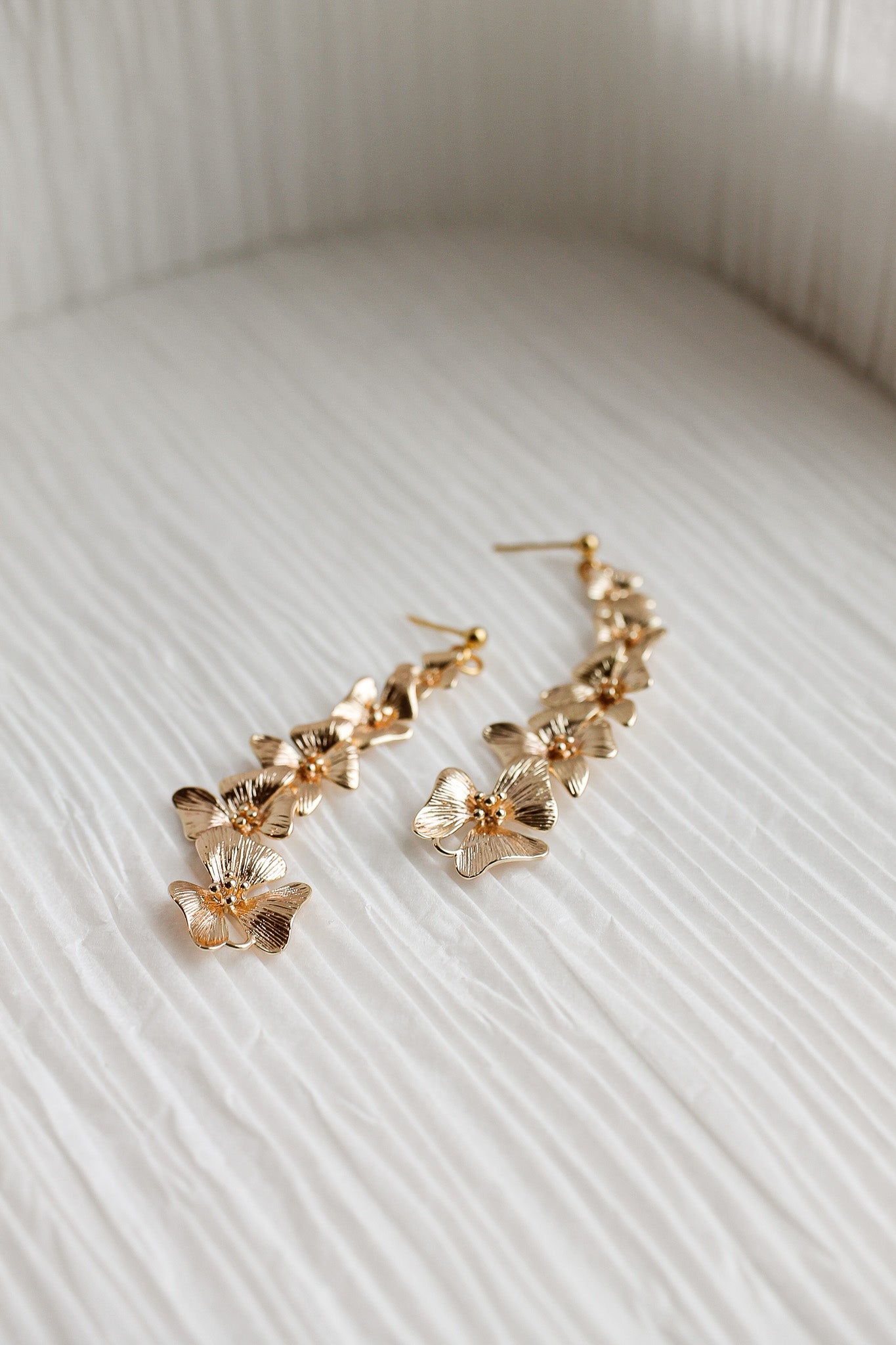 Gold floral earrings are displayed, with one longer piece featuring multiple petals and a shorter piece nearby. They rest on a softly textured, light-colored surface.