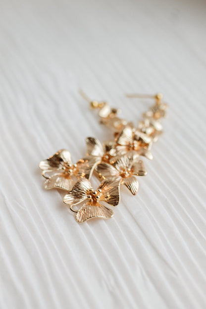 Gold floral earrings rest on a textured white surface, showcasing their intricate design. The flowers feature detailed petals and are arranged in a cascading pattern, suggesting elegance.