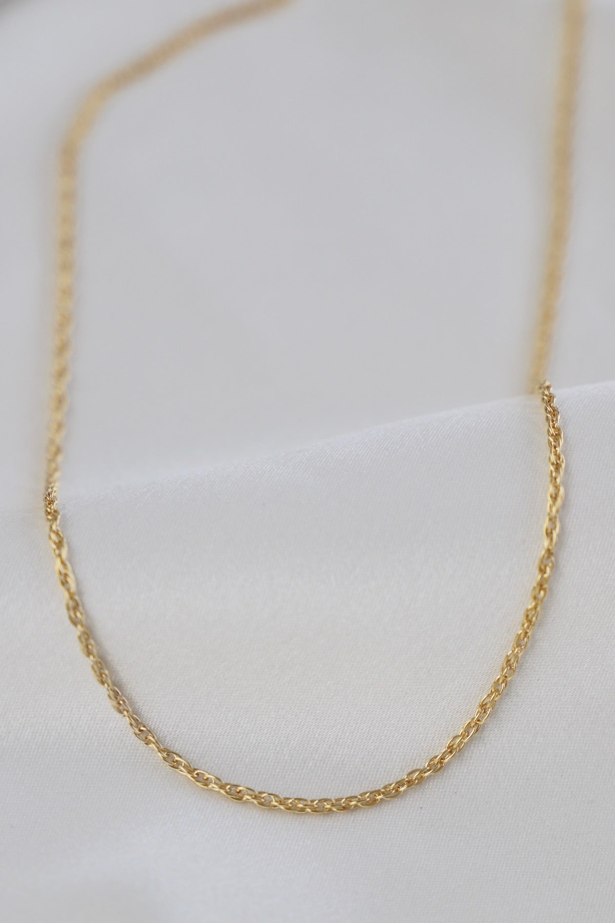 A delicate gold chain rests atop smooth, white fabric. The chain features a twisted design, reflecting light softly, suggesting elegance and sophistication in its simple presentation.