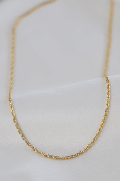 A delicate gold chain rests atop smooth, white fabric. The chain features a twisted design, reflecting light softly, suggesting elegance and sophistication in its simple presentation.