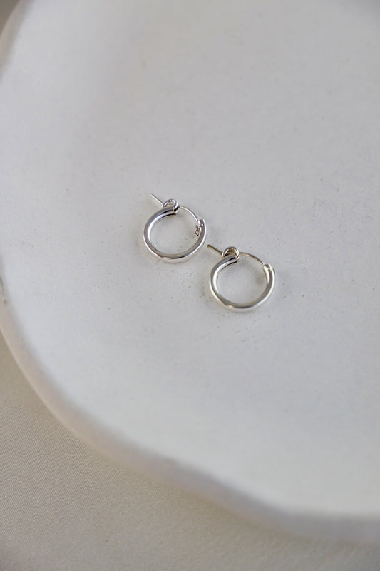 Two silver hoop earrings rest on a smooth, pale ceramic dish. The background is a soft, neutral fabric, creating a minimalist and elegant presentation.