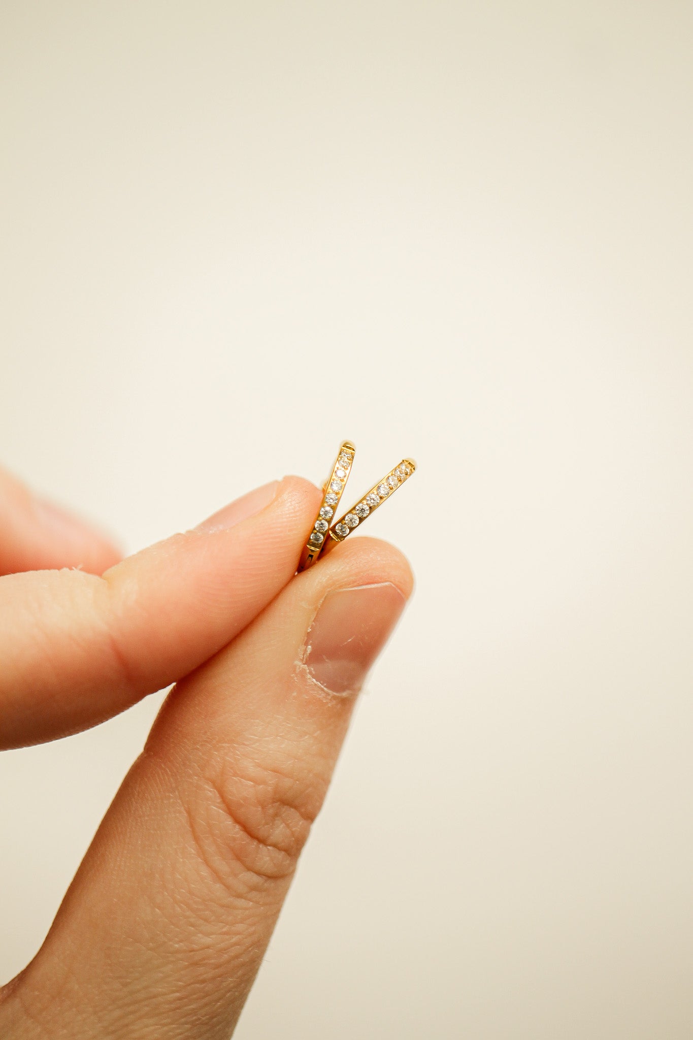 A hand holds two delicate gold rings adorned with small gemstones. They are positioned closely together against a light, neutral background, emphasizing their intricate design and sparkle.