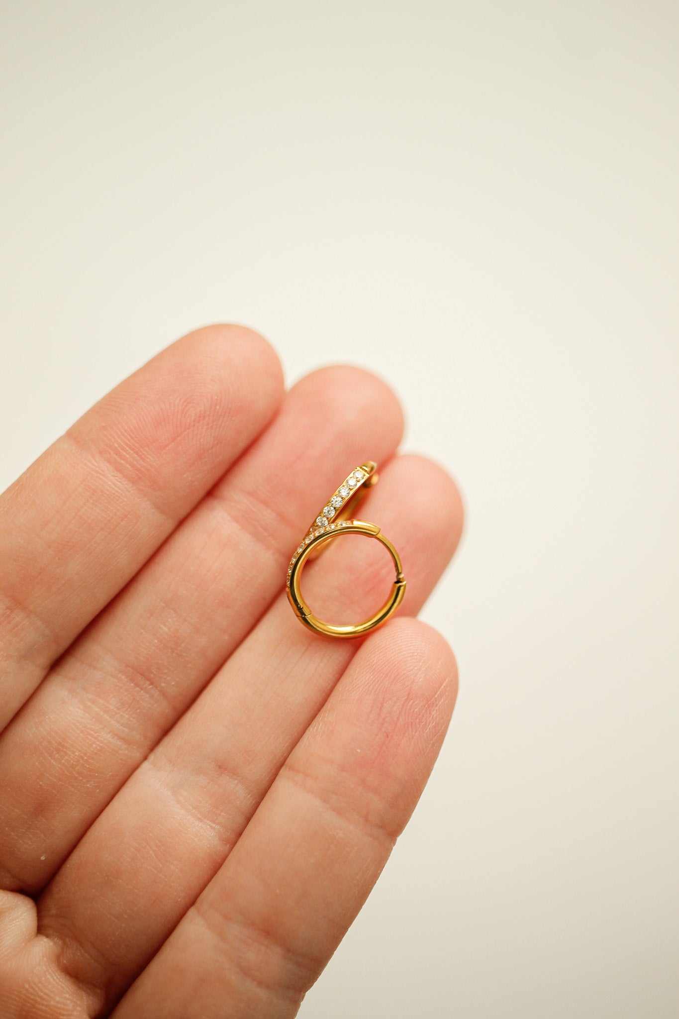 A gold hoop earring, adorned with small sparkling stones, is held gently between fingers against a soft, light-colored background. The design features a curved shape, showcasing elegance.