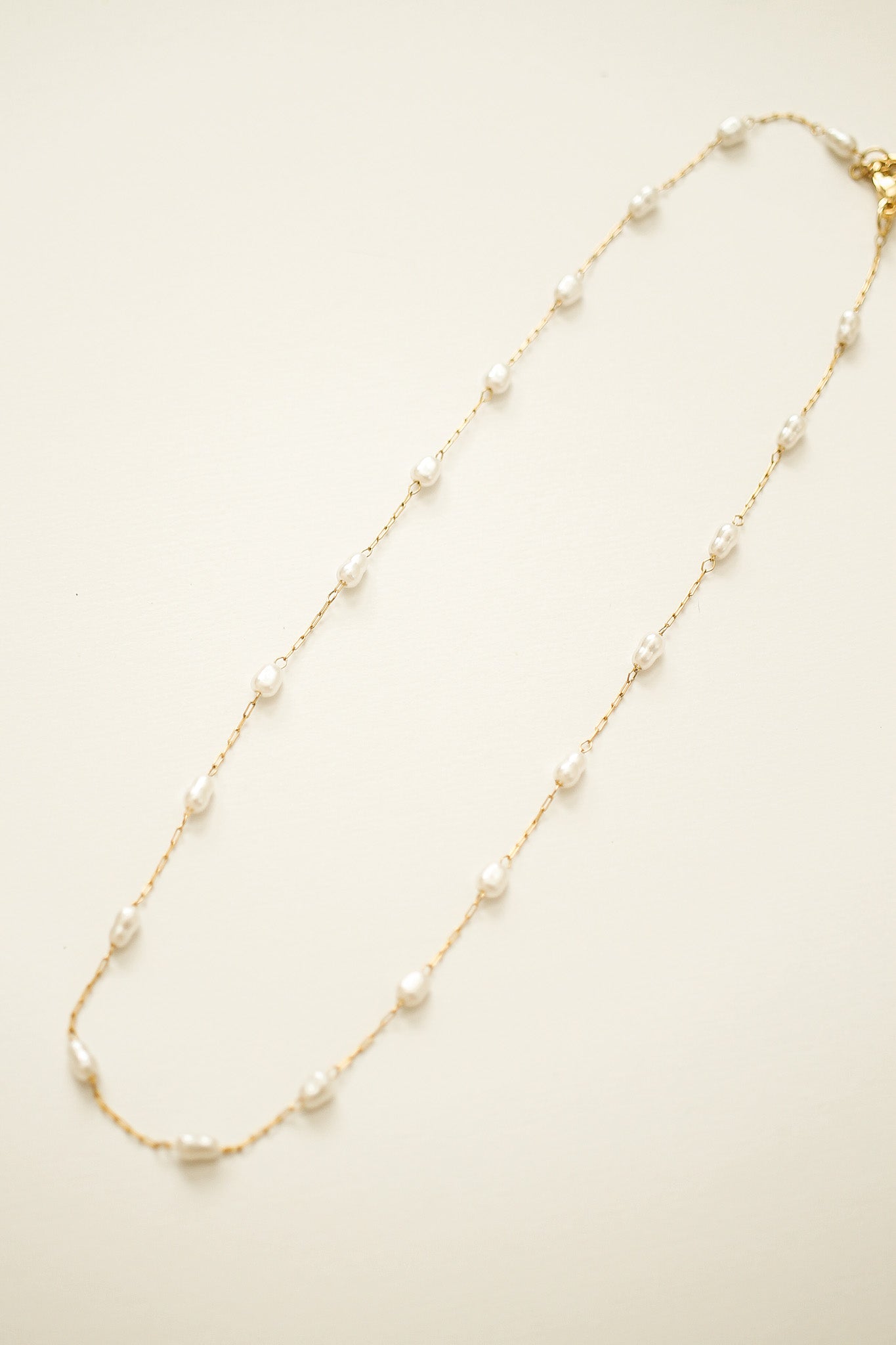 A delicate gold chain necklace features evenly spaced small white pearls. It is laid flat against a soft, light-colored background, emphasizing its elegance and simplicity.