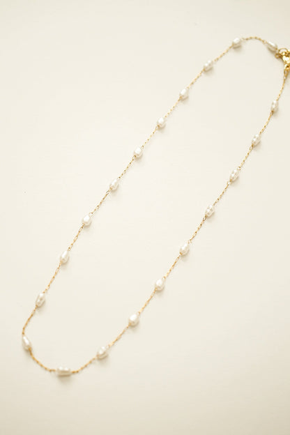 A delicate gold chain necklace features evenly spaced small white pearls. It is laid flat against a soft, light-colored background, emphasizing its elegance and simplicity.