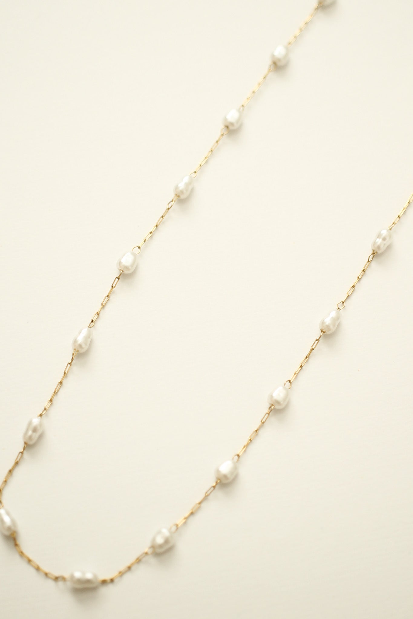 A delicate gold chain features irregularly shaped white pearls spaced along its length, elegantly lying flat against a soft, neutral background.