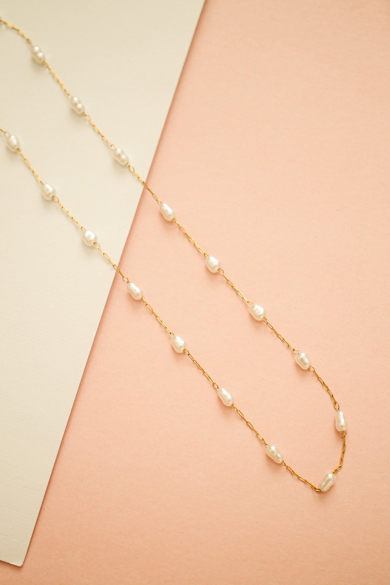 A gold chain necklace with irregularly shaped white pearls is laid out elegantly against a soft pink background, accompanied by a light-colored paper card.