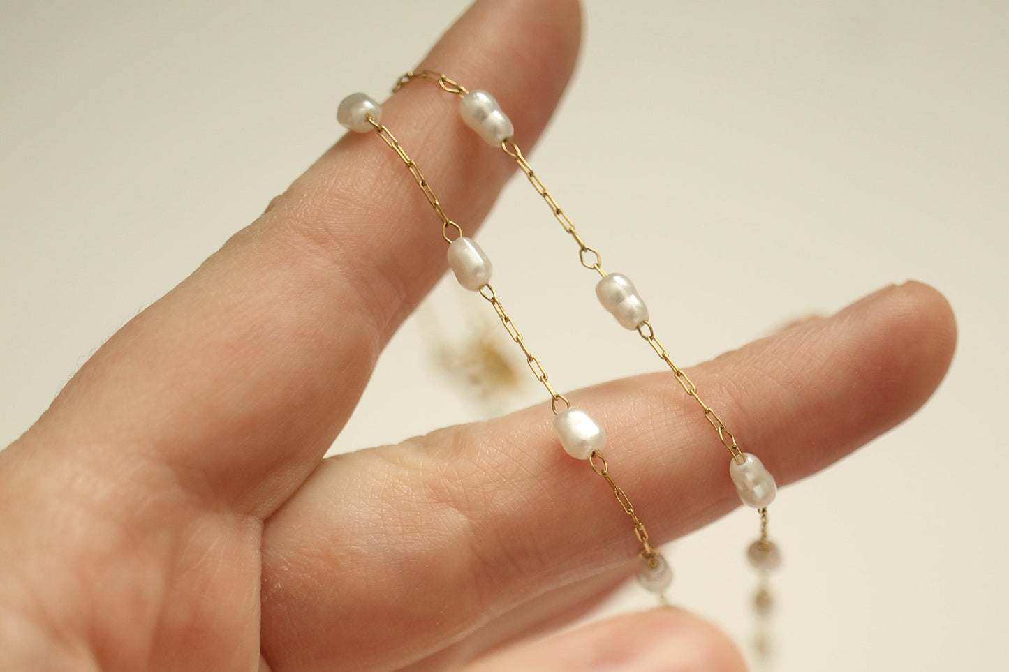 A hand holds a delicate chain adorned with irregularly shaped pearls, showcasing its elegance against a softly lit, neutral background. The gold links connect the pearls, highlighting their sheen.