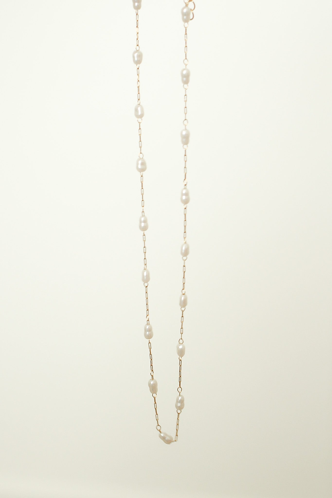 A delicate pearl necklace hangs vertically, with evenly spaced pearls linked by fine gold chain, against a soft, light-colored background, highlighting its elegance and simplicity.