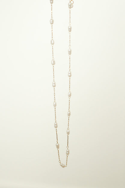 A delicate pearl necklace hangs vertically, with evenly spaced pearls linked by fine gold chain, against a soft, light-colored background, highlighting its elegance and simplicity.