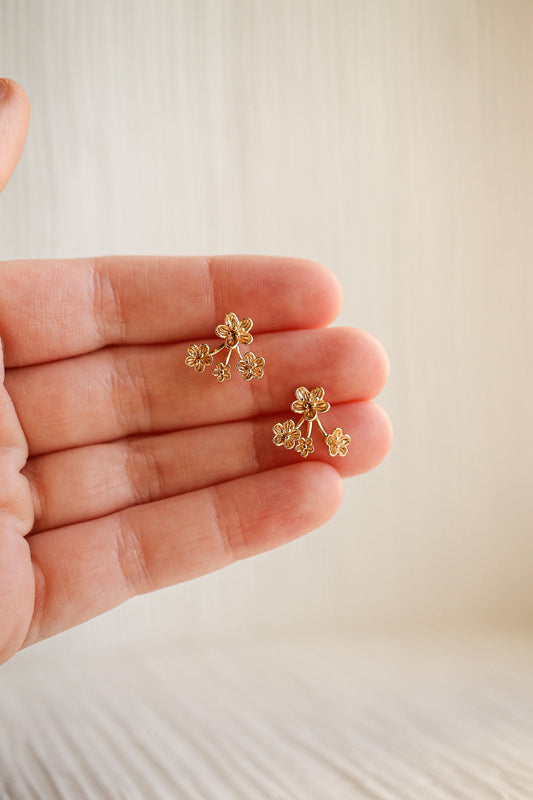 A hand holds a pair of delicate, gold floral earrings, showcasing their intricate design against a soft, neutral background, highlighting both the jewelry and the hand’s graceful gesture.