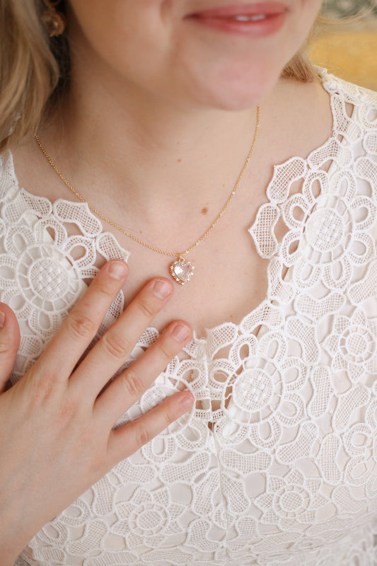 A delicate gold necklace with a heart-shaped pendant is being gently touched by a woman wearing a white lace top, highlighting the jewelry against a soft, elegant background.