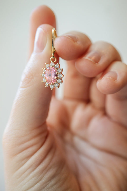 A hand holds a pink gemstone earring adorned with sparkling gemstones. The background is soft and blurred, suggesting an indoor setting, emphasizing the earring's detail and craftsmanship.