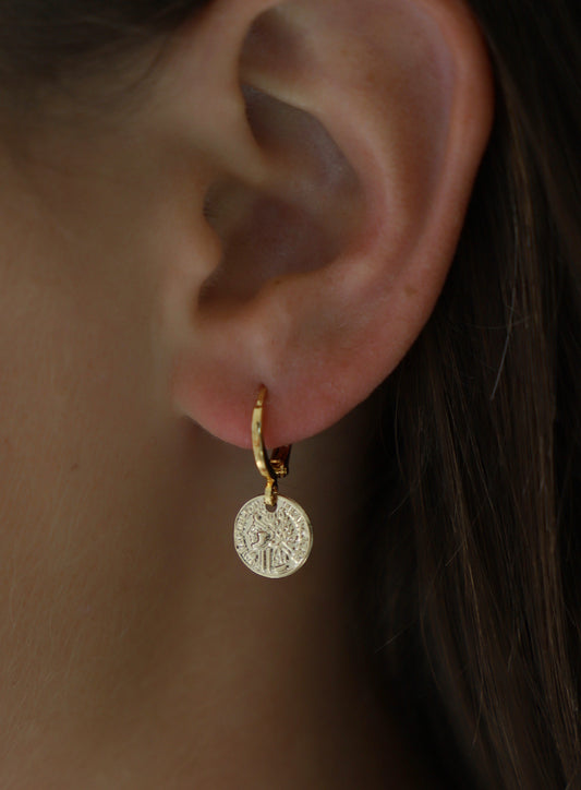 Gold hoop earring featuring a circular, intricately detailed pendant hangs from a person's ear. The backdrop is slightly blurred, emphasizing the earring's design and the ear's natural texture.