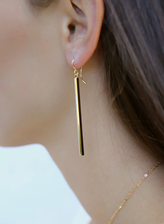 A gold dangle earring, featuring a slim, vertical bar, hangs elegantly from the lobe of a woman's ear, partially visible hair and skin serve as a neutral backdrop.