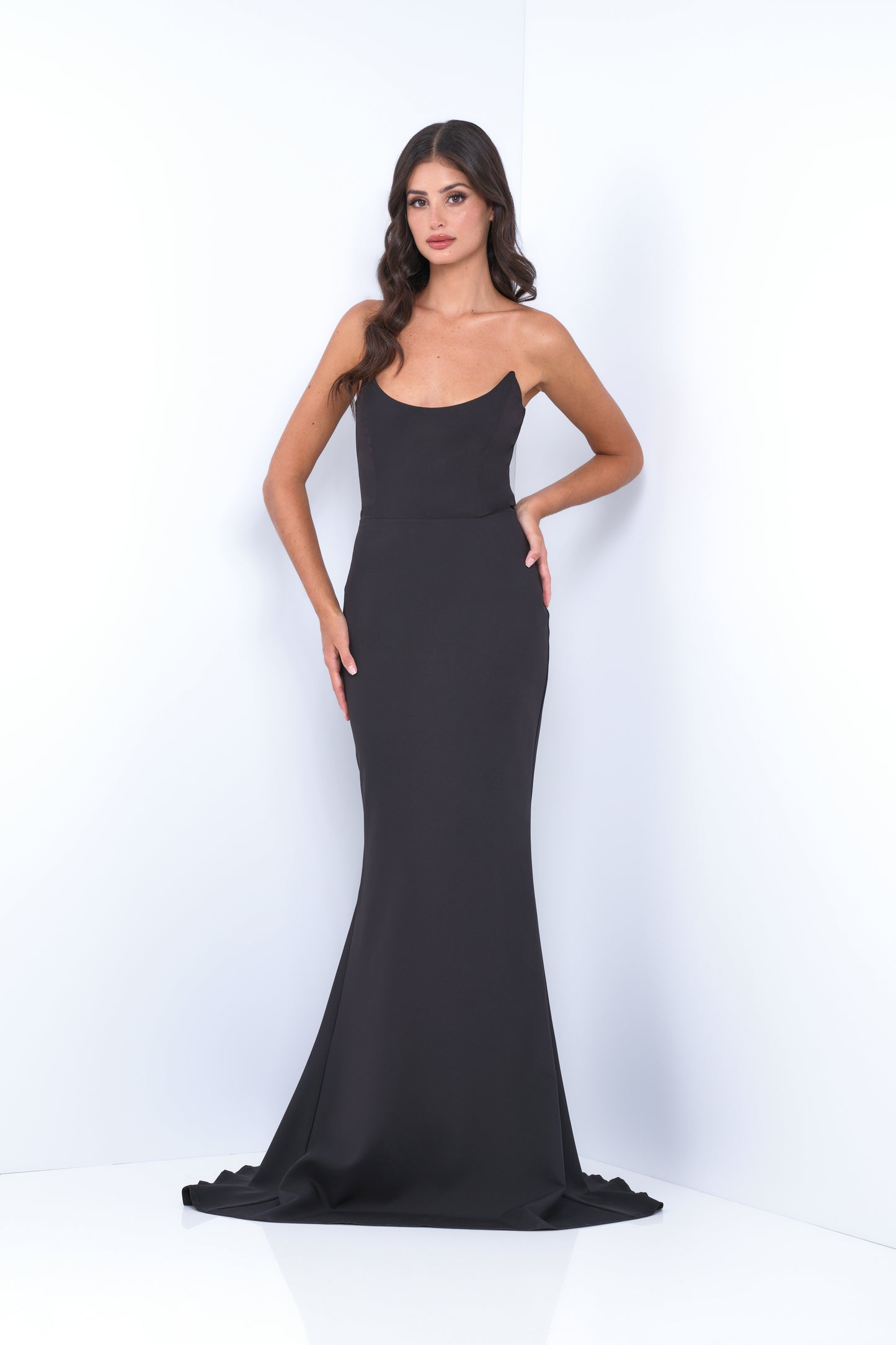 A woman stands elegantly in a form-fitting black gown, with a flare at the bottom. She poses against a plain white backdrop, showcasing the dress's sleek silhouette.