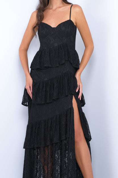 A woman is posing, showcasing a black lace dress with layered ruffles and a thigh-high slit. The background is light and neutral, highlighting the dress's details.