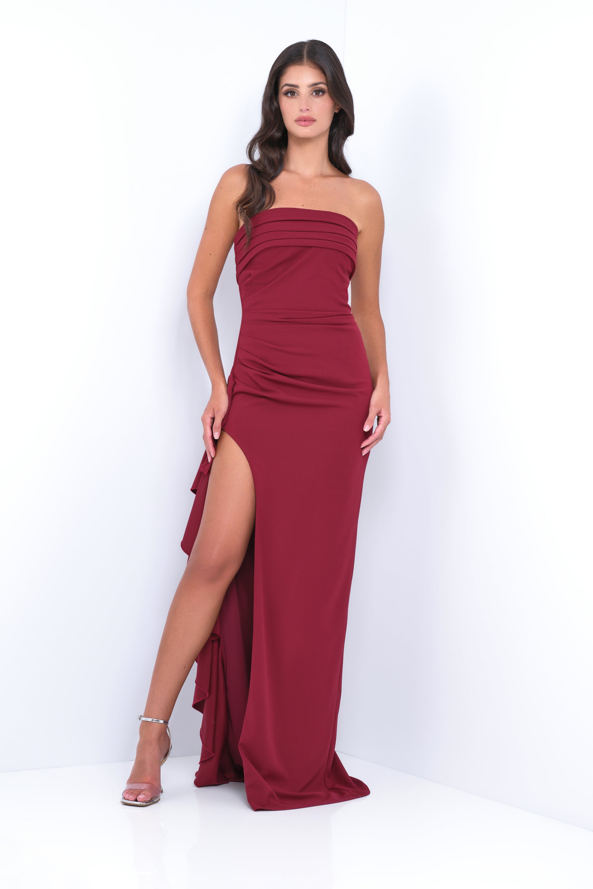 A woman stands confidently in a fitted, burgundy evening gown with a thigh-high slit. She poses against a bright, white background, showcasing the sleek design of her attire.