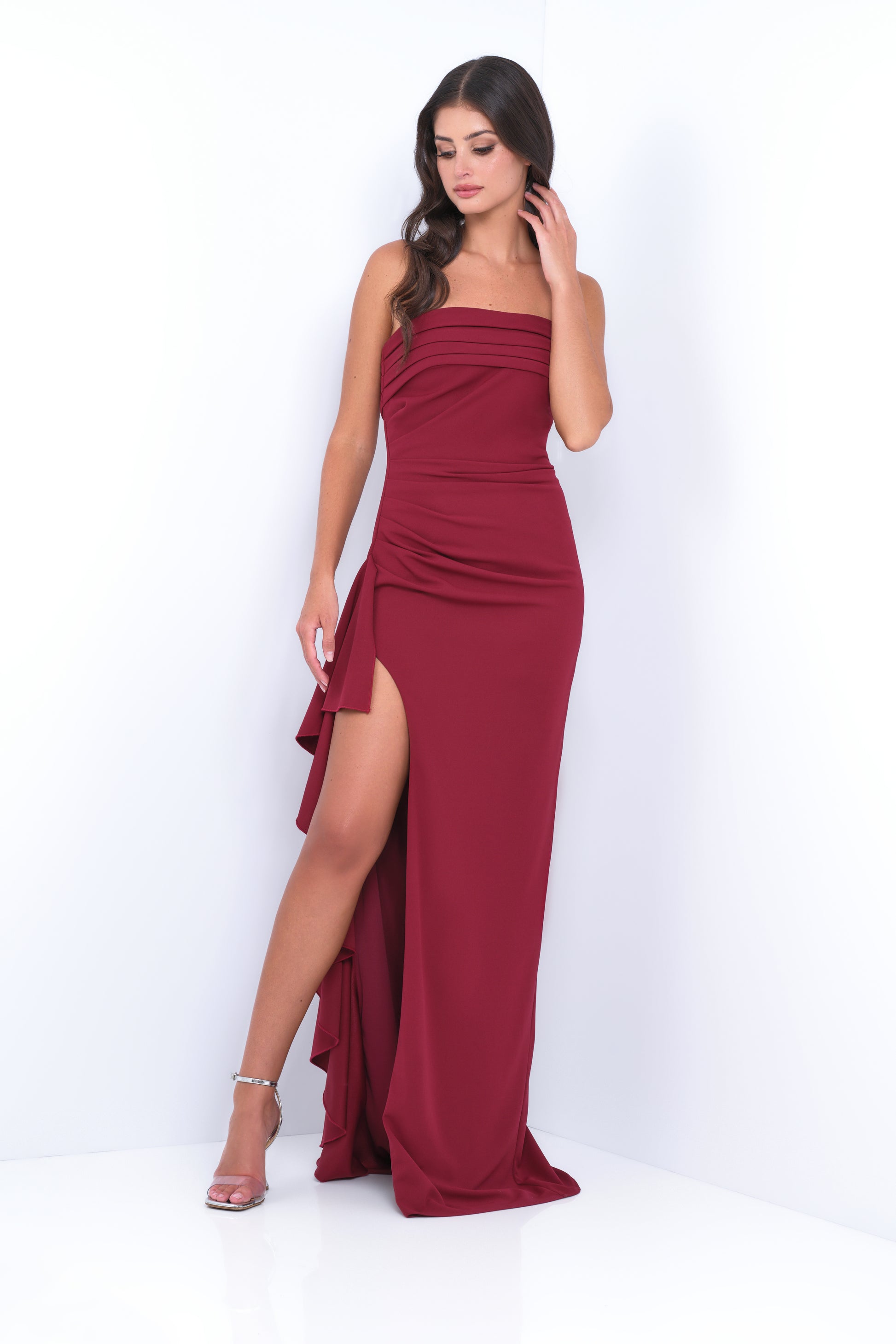 A woman stands elegantly in a burgundy evening gown with a thigh-high slit, striking a poised pose against a minimalist white backdrop, exuding confidence and grace.