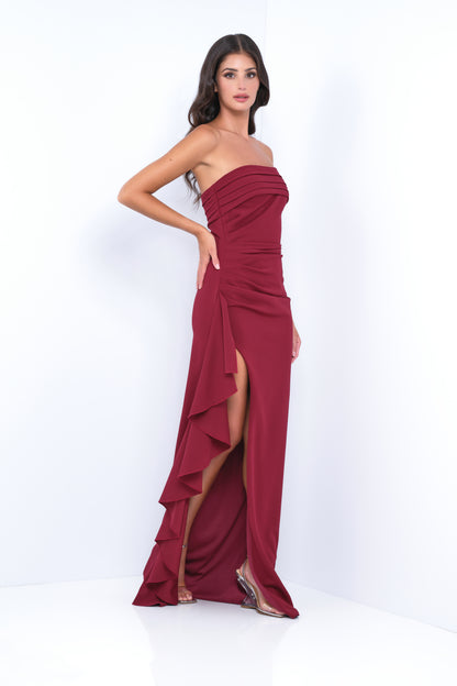 A person stands confidently in a burgundy strapless dress with pleats and a high slit, showcasing elegant ruffles. The background is minimalistic and white, enhancing the outfit's prominence.