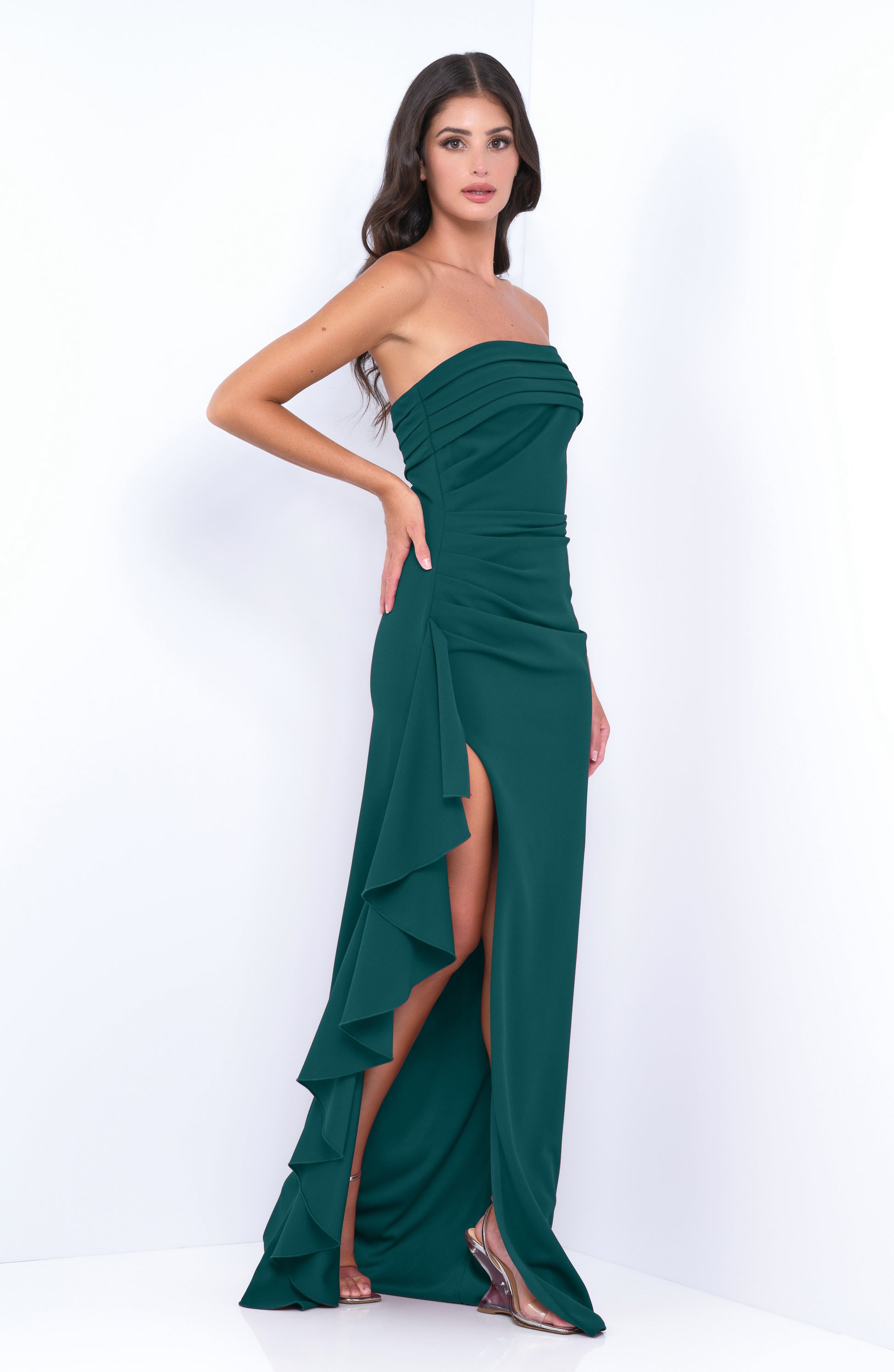 A woman stands confidently in a strapless, emerald green gown with side ruffles and a thigh-high slit, set against a minimalistic white background, highlighting her elegant pose and attire.