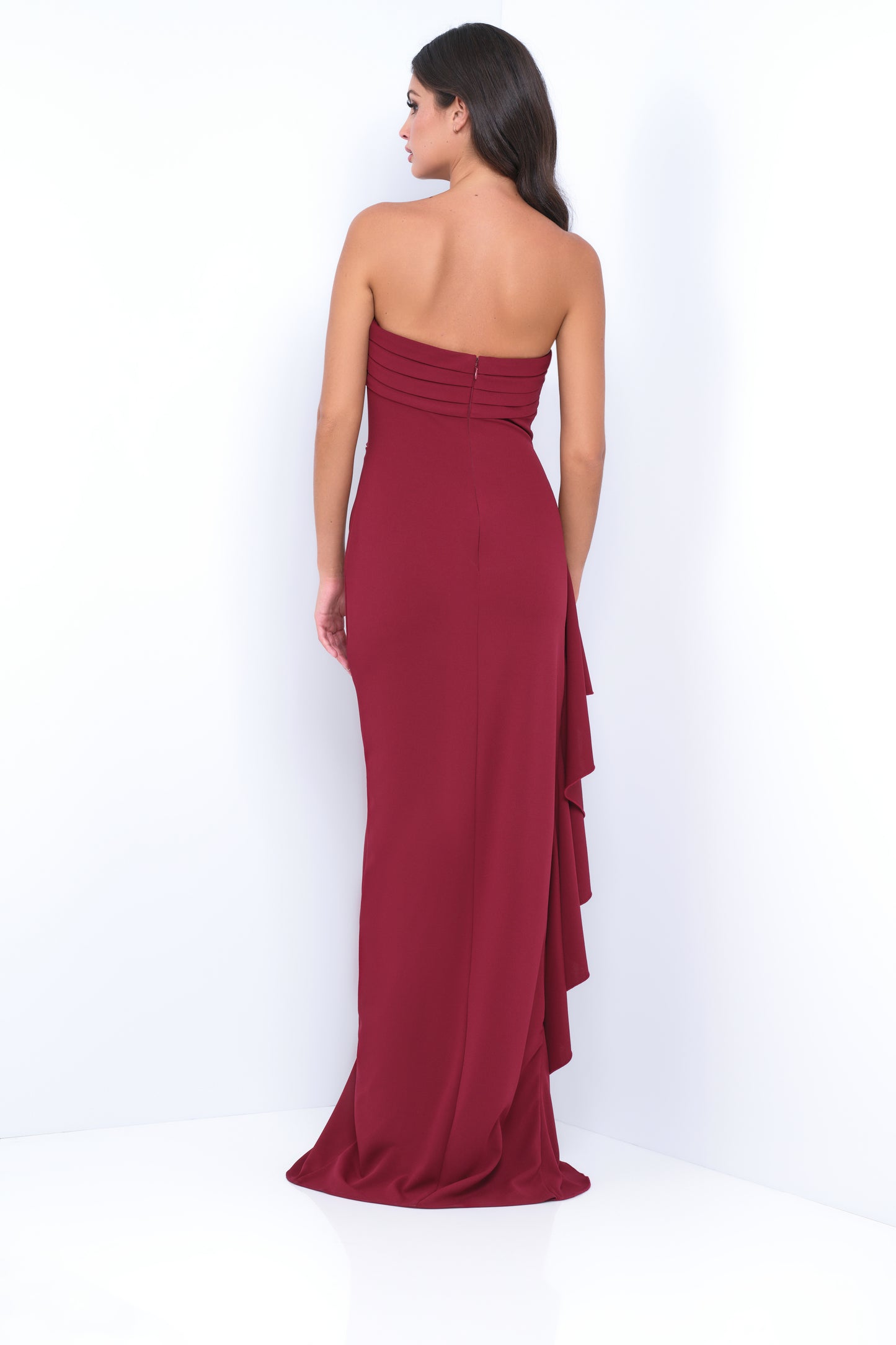 A woman stands in a fitted, strapless burgundy gown with layered draping at the back, showcasing the dress's elegant design against a plain white background.