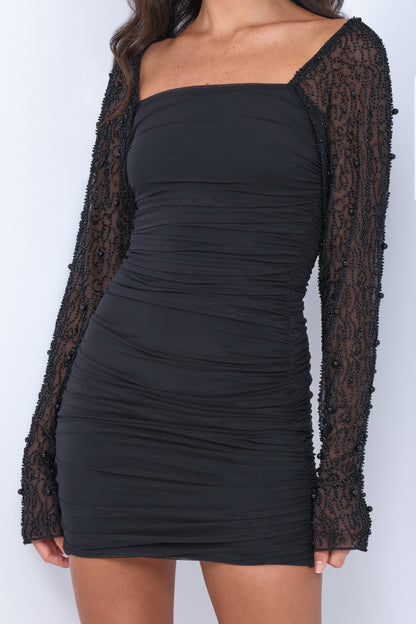 A black dress with a fitted body and ruched fabric features long, sheer sleeves adorned with intricate beadwork. The model stands against a neutral background, showcasing the elegant design.