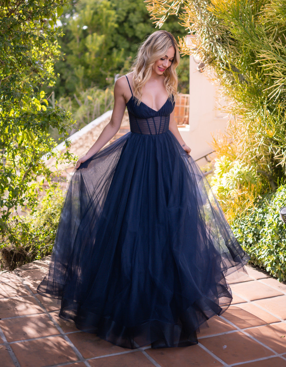 A person wears a flowing navy gown with a fitted bodice and full skirt, gracefully looking down as they stand outdoors among lush greenery and tiled pathways.