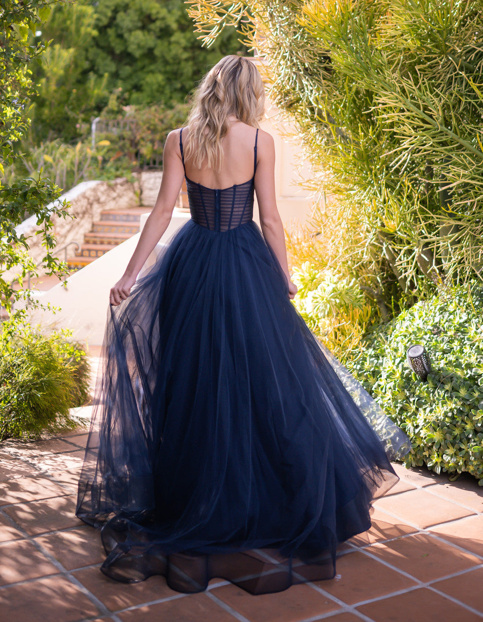 A woman in a navy gown with a sheer skirt walks away, revealing a lush garden and stone steps in bright, natural light, creating an elegant outdoor setting.