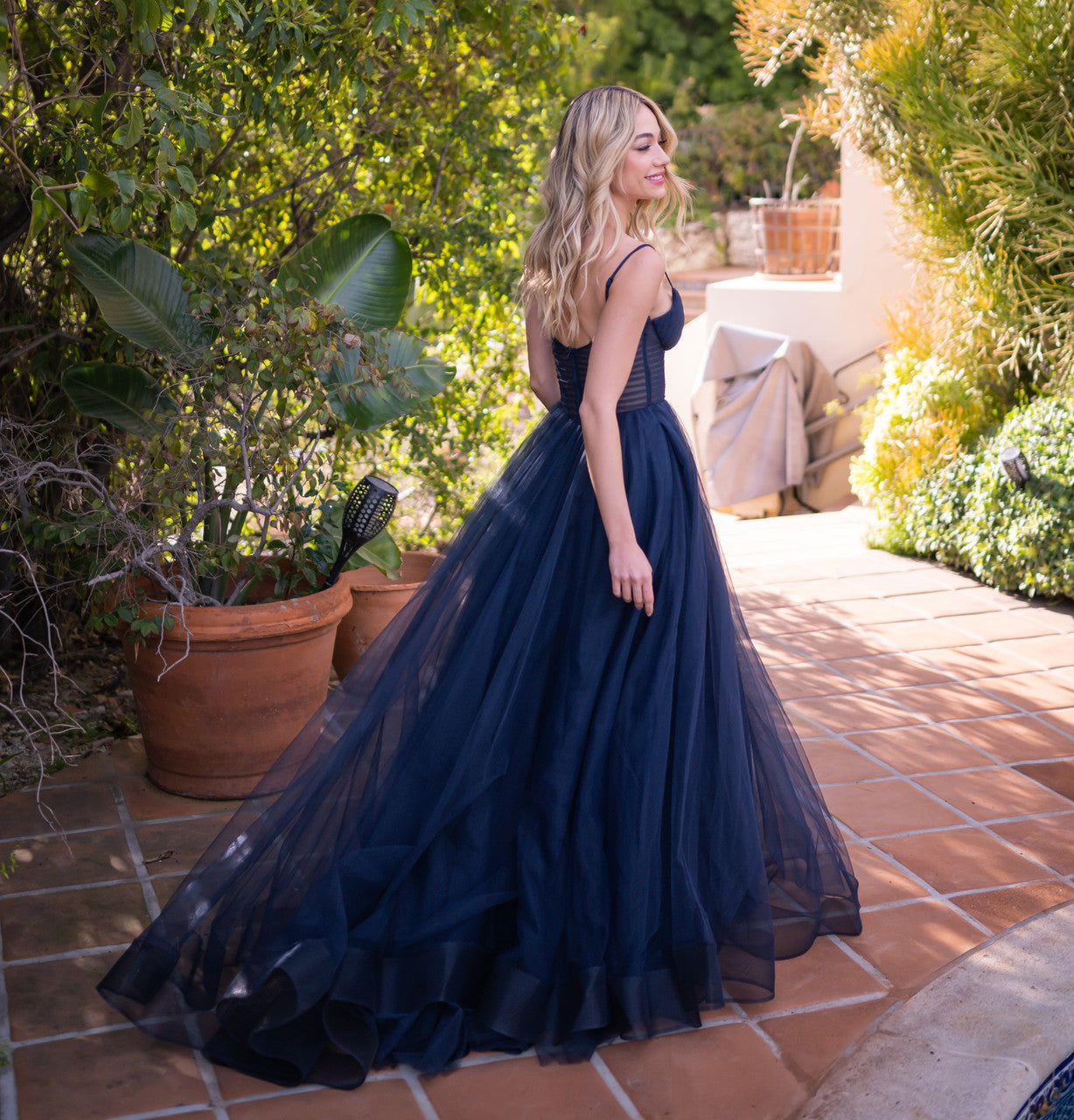 A woman in a flowing navy gown turns gracefully, showcasing the dress's layers as she walks along a terracotta pathway surrounded by greenery and potted plants in a sunny outdoor setting.