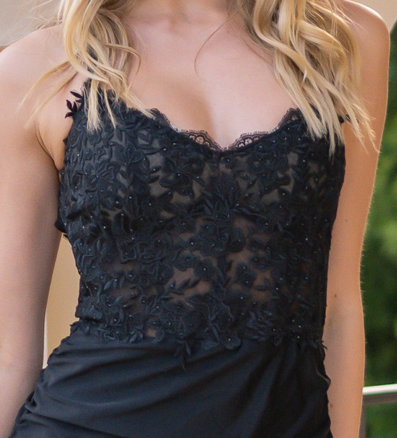 A person wearing a black, embroidered dress poses confidently. The dress features floral patterns and intricate beadwork, situated outdoors with blurred greenery in the background.